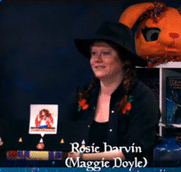 a red-haired woman in a black hat and shirt points behind her and says bang in a very unenthusiastic tone