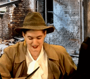 a dark haired person in a fedora and trench coat blinks at someone offscreen and says oh we are gonna have a talk later